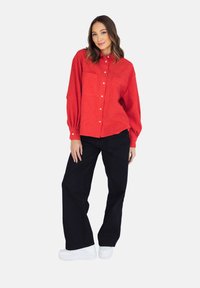 Czerwona, zapinana na guziki koszula o luźnym kroju, z dwoma kieszeniami z przodu i podwiniętymi rękawami, w połączeniu z czarnymi spodniami na szerokich nogawkach i białymi butami na platformie.