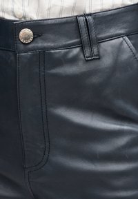 Gros plan sur la ceinture d'un pantalon en cuir noir avec un bouton argenté et des coutures, porté avec une chemise blanche à rayures.