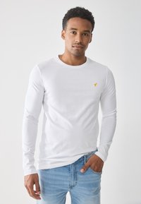 Camisa blanca de manga larga en una tela suave, con un pequeño logo amarillo en el lado izquierdo del pecho. Combinada con unos vaqueros de mezclilla azul claro.