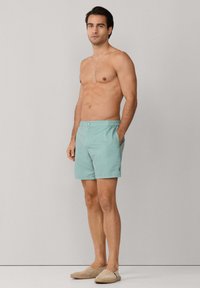 Hombre sin camisa posando contra un fondo liso, lleva pantalones cortos de baño color teal claro y zapatos beige sin cordones, con las manos en los bolsillos, mirando hacia adelante.