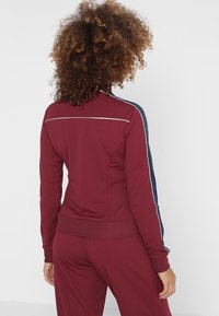 Bordeauxfarbene Sportjacke mit marineblauen Seitenstreifen, taillierter Schnitt und geripptem Saum. Glattes Material mit weißen Nähten auf der Rückseite.