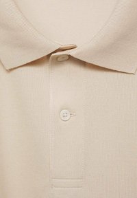 Beige polo triko s vroubkovaným límcem, dvoubuttonovou légu a texturovanou pleteninou. Tkanina se zdá být měkká a mírně pružná.
