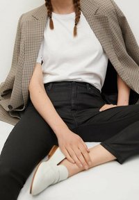 Jeans negros de talle alto, camiseta blanca ajustada y una chaqueta de cuadros marrón. Zapatillas blancas de lona con suela de madera. Modelo sentado de manera casual.