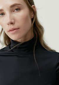 Top negro de cuello alto hecho de una tela lisa, con un diseño ajustado y un leve brillo. El cabello es ondulado, cubriendo parcialmente el hombro.