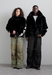 Chaquetas de pelo sintético negro con cuellos amplios; una de ellas tiene acentos manchados en blanco. Los pantalones del modelo son de color verde oliva y negro, de corte holgado y con textura.
