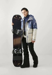 Sniego lenta su blizgiu juodu paviršiumi ir tvirtinimais. Modelis mūvi mėlyną ir grietinėlės spalvos striukę su gėlių raštais, juodas kelnes ir žieminius batus.