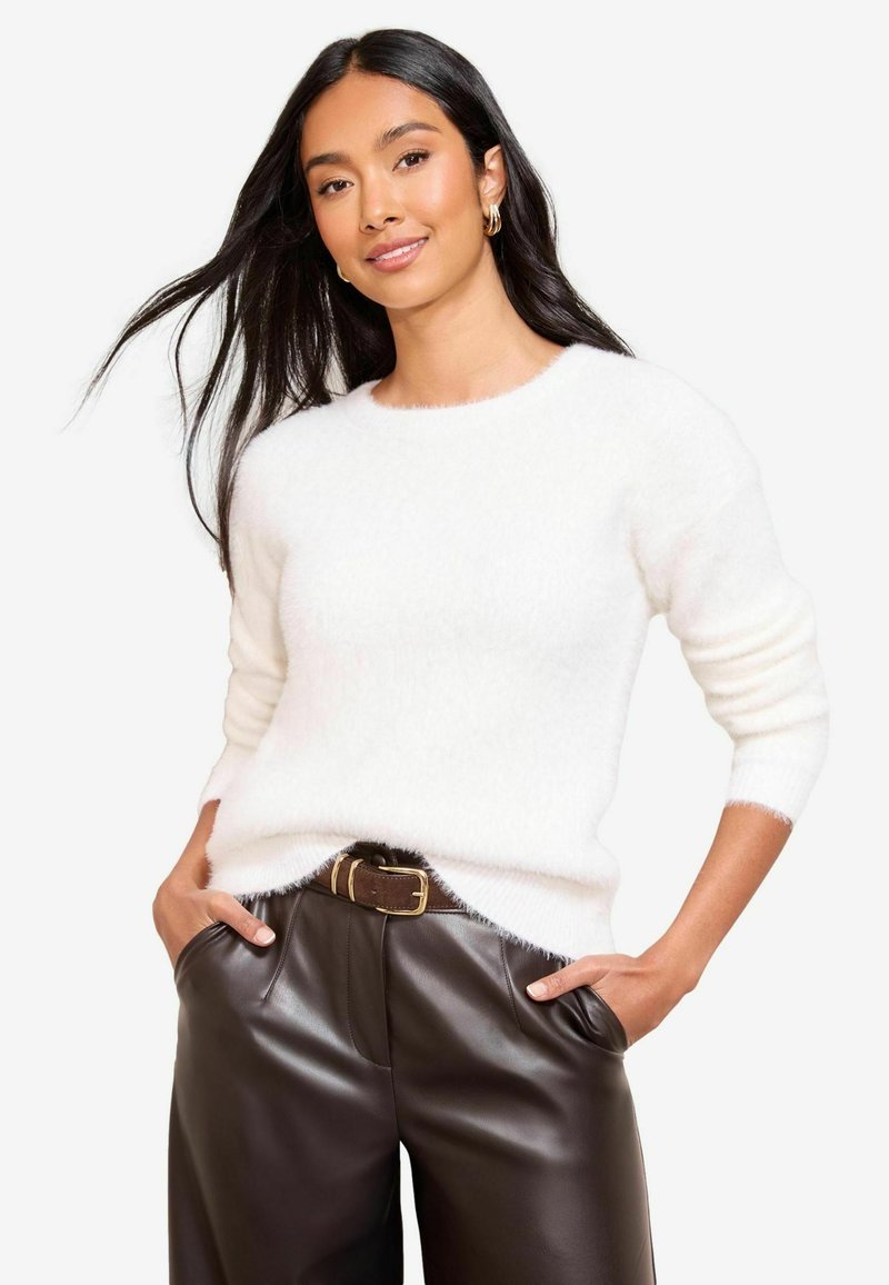 Weißer, flauschiger Pullover mit Rundhalsausschnitt und langen Ärmeln, kombiniert mit dunkelbraunen Lederhosen und einem braunen Gürtel mit goldener Schnalle.