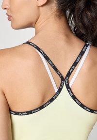 Zwart-witte merklussen kruisen aan de achterkant, bevestigd aan een lichtgele tanktop van gladde stof.