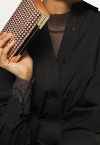 Metaliczna kopertówka w kolorze różowego złota z teksturowanymi ozdobami z kryształków, o prostokątnym kształcie i eleganckim designie, trzymana przez osobę w czarnej koszuli.