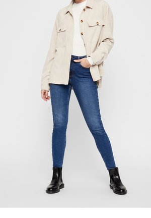 Personne portant une chemise beige boutonnée, un haut blanc, un jean skinny bleu et des bottines noires, debout avec une main dans la poche.