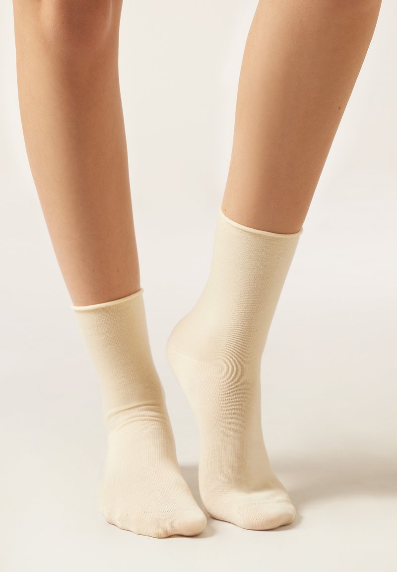 Gambe che indossano un paio di calzini alla caviglia color crema semplici su uno sfondo neutro.