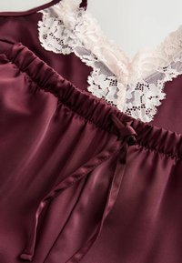 Burgunderfarbene Satin-Camisole mit V-Ausschnitt, Spitzenbesatz und einem elastischen Bund mit einer Schleifenapplikation am unteren Ende.
