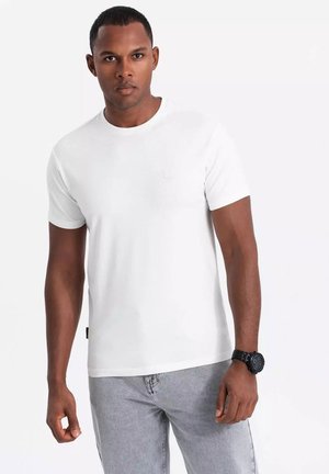 Weißes Baumwoll-T-Shirt mit rundem Ausschnitt und kurzen Ärmeln, mit einem kleinen Logo auf der Brust. Dazu werden hellgraue Jeans getragen.