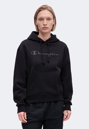 Ung person med kort blondt hår iført en sort Champion hoodie og sorte bukser, stående mod en hvid baggrund.