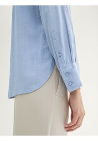 Camicia a maniche lunghe celeste con orlo arrotondato, polsini con bottoni e una morbida texture; abbinata a pantaloni beige.