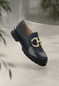 Zwarte leren mocassin met dikke zool en grote gouden decoratieve gesp, zwevend boven een betonnen oppervlak met een vervaagde achtergrond.