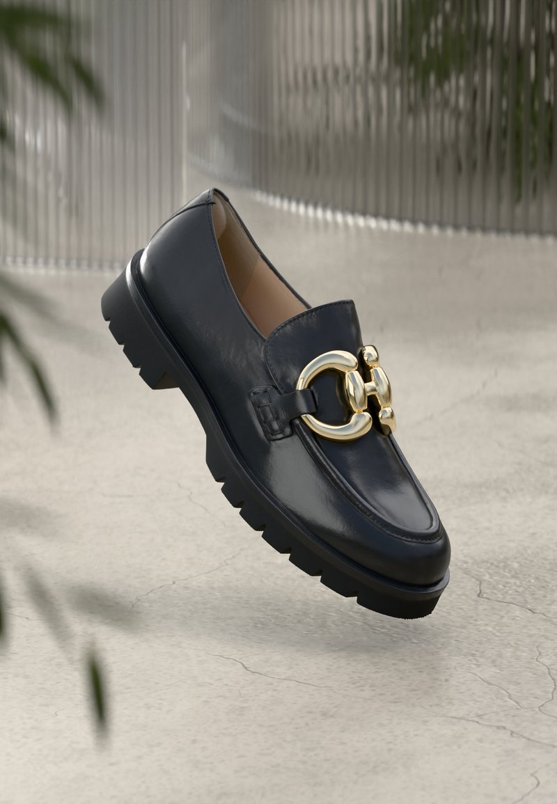 Melnas ādas loafer kurpes ar masīvu zoli un lielu zelta dekoratīvu sprādzi, kas karājas virs betona virsmas ar izplūdušu fona.