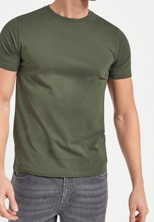 Homme portant un t-shirt moulant à manches courtes vert olive et un jean gris, debout les bras détendus le long du corps devant un fond blanc.