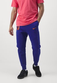 Blaue Nike-Sweatpants mit orangefarbenem Logo und niederländischem Wappen, ausgestattet mit seitlichen Taschen und schmalen Bündchen. Kombiniert mit einem pinken T-Shirt und schwarzen Turnschuhen.