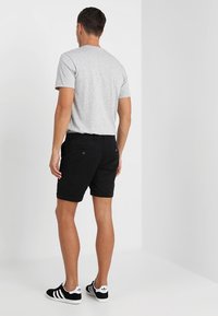 Homme debout de dos portant un t-shirt gris clair, un short noir et des baskets Adidas noires à bandes blanches.