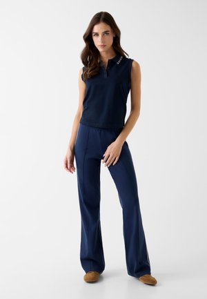 Femeie purtând un tricou polo navy fără mâneci și pantaloni largi asortați, cu pantofi maro, stând pe un fundal alb simplu.