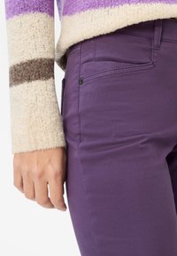 Paarse katoenen broek met een gladde textuur, voorzien van een zijzak en een zwart knoopdetail; gecombineerd met een gestreepte wollen trui.