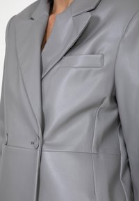 Blazer grigio in similpelle con revers a punta, taschino sul petto e chiusura a doppio bottone. Texture liscia e design strutturato.