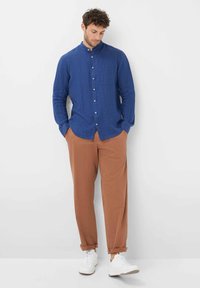 Camicia azzurra in lino a maniche lunghe con chiusura a bottoni e taschino sul petto, abbinata a pantaloni color ruggine e sneaker bianche.