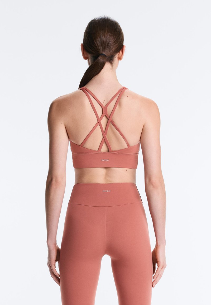 Ženska z rjavimi lasmi v čopku, oblečena v rožnato športno modrček s prekrižanimi naramnicami in ujemajoče visokopasne pajkice, prikazana od zadaj.