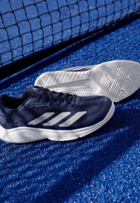 Granatowe buty sportowe z białymi akcentami; syntetyczna cholewka, siateczkowe detale, teksturowana gumowa podeszwa z bieżnikiem, umieszczone na niebieskiej nawierzchni.