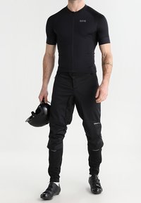 Jersey de ciclismo negro de manga corta y pantalones; fabricados en tejido elástico. Incluye una cremallera y un casco negro sostenido en una mano.