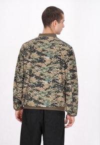 Jakken med camouflagemønster i beige, grøn og sort har lange ærmer, en rund krave og elastiske manchetter, og er parret med sorte bukser.