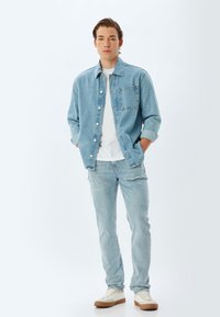 Una chaqueta de denim azul claro con un bolsillo en el pecho, llevada sobre una camiseta blanca. Combinada con unos jeans azul claro y zapatillas crema con suelas marrones.