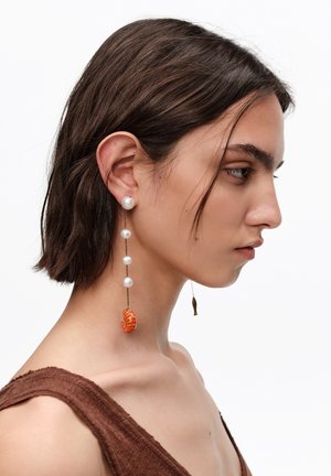 Kobieta o krótkich ciemnych włosach, nosząca długą zwisającą kolczyk z białych pereł i pomarańczowym muszelkowym zawieszkiem, ubrana w brązowy top bez rękawów.