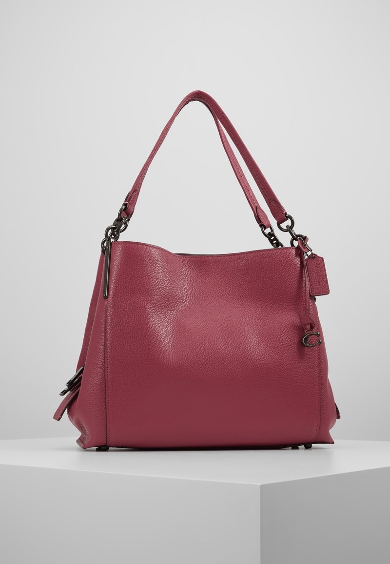 Grand sac à main en cuir rose texturé avec double poignée, métal et petite étiquette attachée, posé sur un socle blanc sur fond gris.