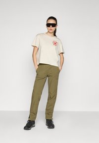Beige t-shirt met korte mouwen en bloemenborduursel, gecombineerd met olijfgroene tapered broeken en zwarte veterschoenen, voor een effen achtergrond.