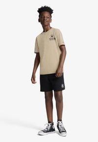 Μπεζ βαμβακερό μπλουζάκι με κοντά μανίκια και μαύρο γραφικό σχέδιο στο στήθος. Φορεμένο με μαύρο σορτς και μαύρα ψηλά αθλητικά παπούτσια.