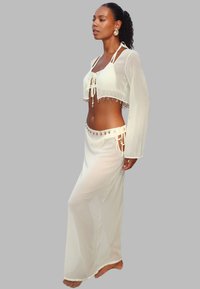 Femme portant un ensemble deux pièces transparent blanc avec des détails en coquillage, debout pieds nus contre un fond gris uni dans une pose de profil.