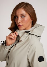 Lichtgrijze waterdichte jas met een hoge kraag, verstelbaar koord, zwarte accenten en een logo-opdruk op de mouw. Zachte textuur.