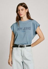 Mėlynas laisvas tinkantis marškinėliai su trumpomis rankovėmis, kuriose puikuojasi „PEPE JEANS LONDON“ kontrastingame dizajne, derinami su šviesiai mėlynomis džinsomis ir juodu diržu.
