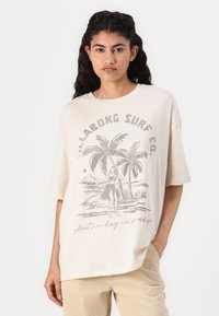 T-shirt oversize de couleur crème en tissu doux, avec un imprimé graphique de palmiers et d'une danseuse hula, accompagné d'accents de texte gris.