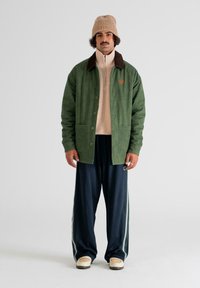 Grøn fløjlsjakke med brun krave, beige zip-up sweater under, mørkeblå bukser med sidelister og beige sko. Strikket beige beanie.