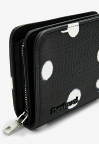 Schwarze Geldbörse mit weißen Punkten, strukturierte Oberfläche, mit Reißverschlussverschluss und schwarzem Logo-Akzent. Kompaktes Design für einfachen Transport.