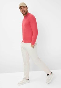 Roter langärmliger Strickpullover mit Rundhalsausschnitt, kombiniert mit hellbeigen Hosen und weißen Sneakers. Der Mann geht lässig.