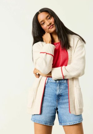 Kobieta z długimi czarnymi włosami, ubrana w czerwoną bluzkę, beżowy kardigan z czerwonym obszyciem oraz dżinsowe szorty, stojąca na tle jednolitego jasnego tła.