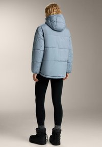 Osoba v světle modré péřové bundě s kapucí, černých legínách, šedých ponožkách a černých zimních botách, stojící a obrácená zády na neutrálním pozadí.