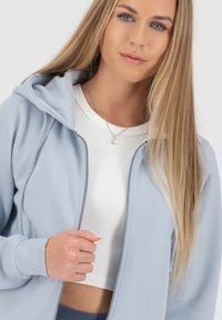 Felpa zip in cielo chiaro in tessuto morbido, con cappuccio e polsini a coste, indossata sopra una maglietta bianca a coste. Design minimalista senza motivi.