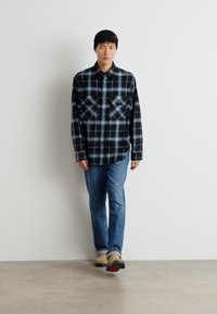 Karirana flanelasta srajca v črni in svetlo modri barvi, skupaj z blue jeans hlačami in bež čevlji. Srajca ima dve žepoma na prsih in gumbasto zasnovo.