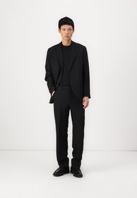 Schwarzer Anzug mit einer maßgeschneiderten Jacke und geraden Hosen, kombiniert mit einem schwarzen T-Shirt und schwarzen Lederschuhen, vor einem weißen Hintergrund.