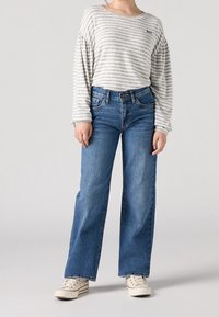 Person som bär en ljusgrå randig långärmad skjorta, blå vida jeans och vita sneakers som står framför en ljus bakgrund.